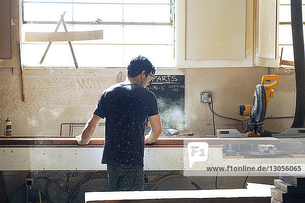 Rückansicht eines männlichen Zimmermanns  der mit einer Elektrosäge in der Werkstatt Holzbohlen schneidet
