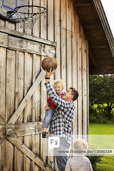 Glücklicher Vater spielt mit Söhnen vor der Scheune Basketball