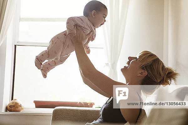 Side view of mother lifting baby girl while sitting on sofa at home