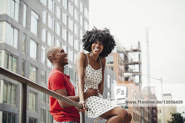 Mann hält Frau fest  die auf einem Geländer vor Gebäuden in der Stadt sitzt