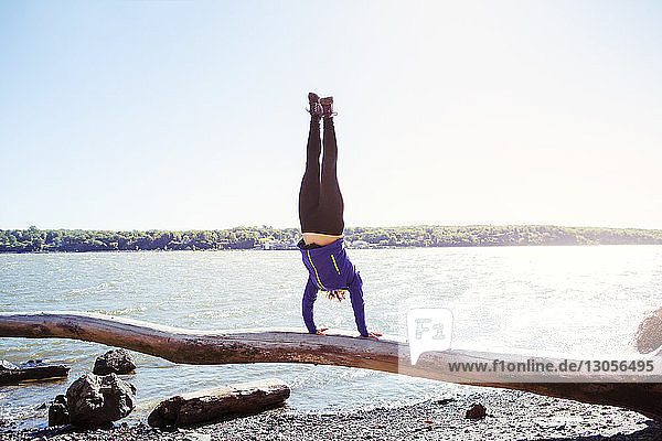 Woman doing handstand on log at riverbank against clear sky