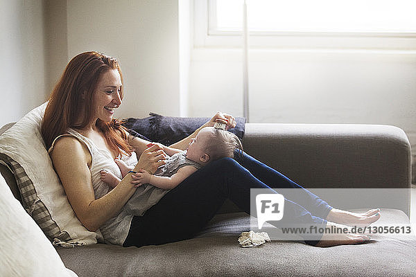 Mother brushing baby girl's hair while sitting on sofa at home