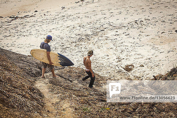 Mann trägt Surfbrett  während er mit einem Freund am Strand spazieren geht