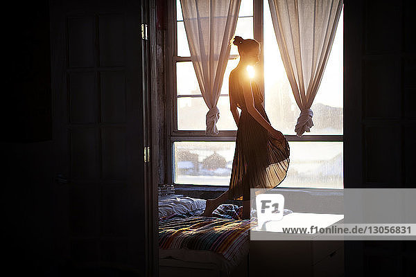 Seitenansicht einer Frau in einem Kleid  die auf dem Bett am Fenster steht und durch die Tür gesehen wird