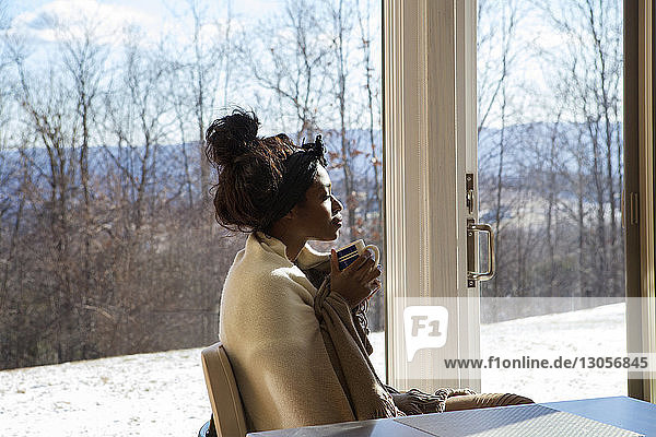 Seitenansicht einer in ein Tuch gehüllten Frau mit geschlossenen Augen  die zu Hause am Fenster sitzt