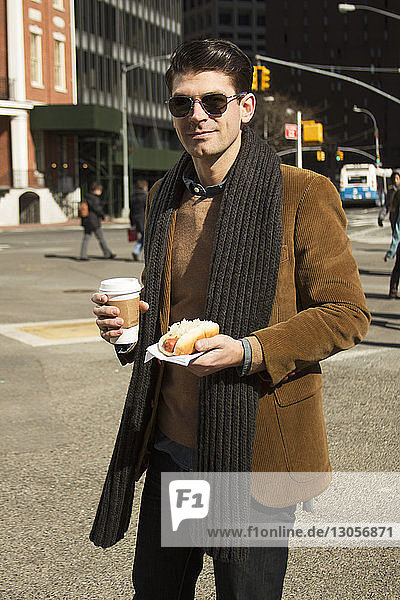 Portrait of man holding coffee and hot dog on city street