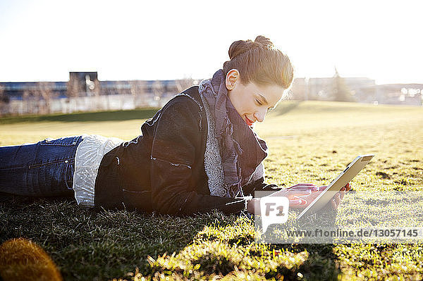 Glückliche Frau benutzt digitales Tablett  während sie auf Grasfeld liegt