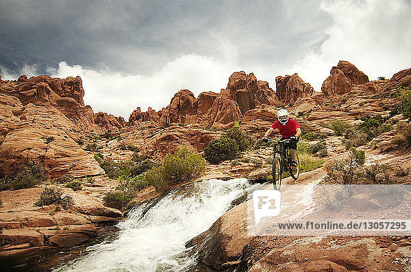 Mountain biker cycling on rocks by river against cloudy sky