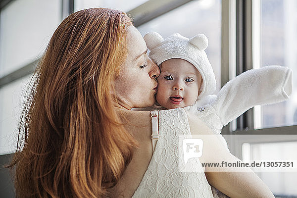 Mother kissing baby girl while standing by window at home