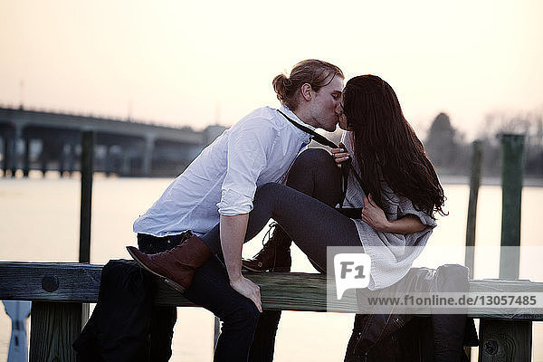 Romantic couple kissing while sitting on railing against sky