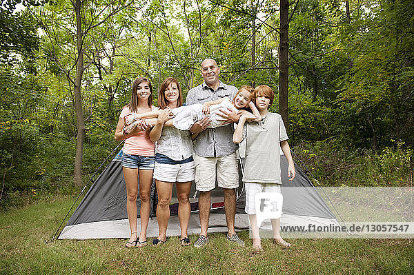Porträt einer glücklichen Familie  die ein Mädchen trägt  während sie im Wald vor einem Zelt steht