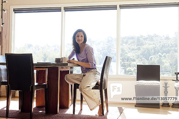 Nachdenkliche Frau hält Kaffeetasse und sitzt zu Hause am Tisch