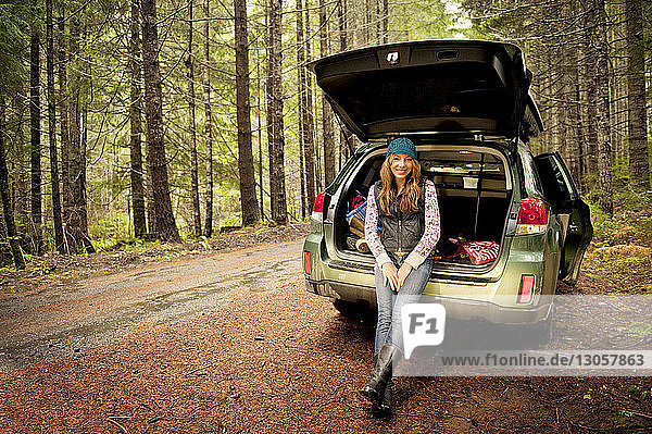 Porträt einer glücklichen Frau  die im Kofferraum eines Autos im Wald sitzt