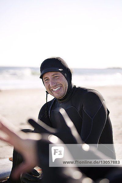 Fröhlicher Surfer im Neoprenanzug  der am Strand am Ufer sitzt