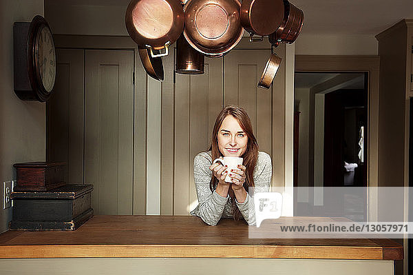 Porträt einer glücklichen Frau  die eine Kaffeetasse hält  während sie sich zu Hause auf eine Holztheke stützt