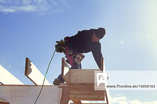 Niedrigwinkelansicht eines Arbeiters mit Bohrmaschine  der auf einem Dachbalken gegen den Himmel steht