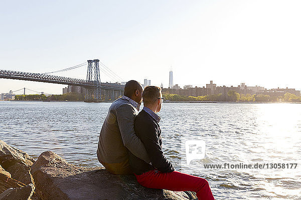 Side view of gay men sitting on rocks while looking at East River on sunny day