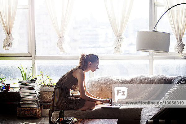 Seitenansicht einer Frau  die einen Laptop-Computer benutzt  während sie zu Hause auf dem Sofa kniend sitzt