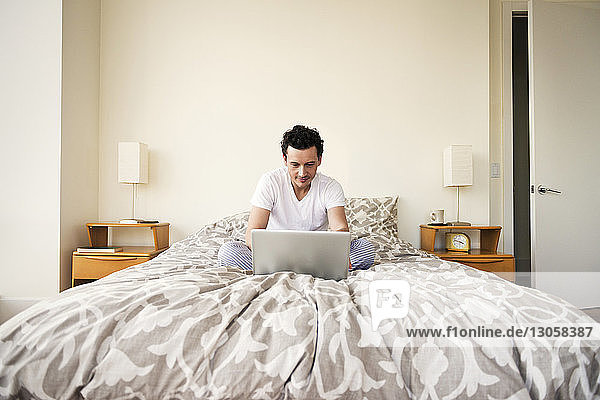 Mann benutzt Laptop  während er zu Hause auf dem Bett sitzt