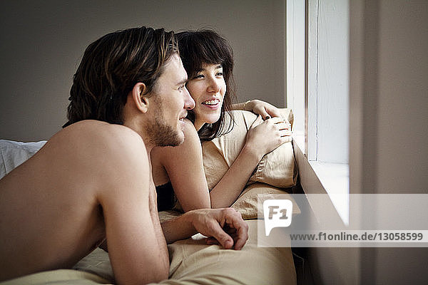 Fröhliches Paar schaut durchs Fenster  während es zu Hause auf dem Bett liegt