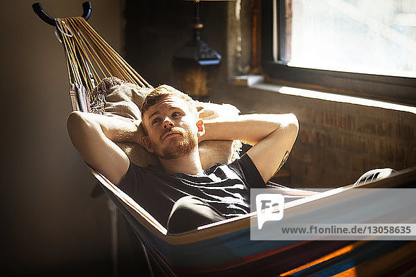 Man with hands behind head looking away while relaxing on hammock at home