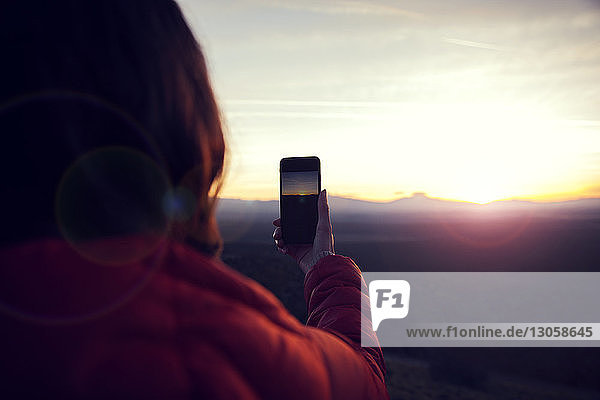Rückansicht einer Frau  die bei Sonnenuntergang Landschaft mit einem Smartphone fotografiert