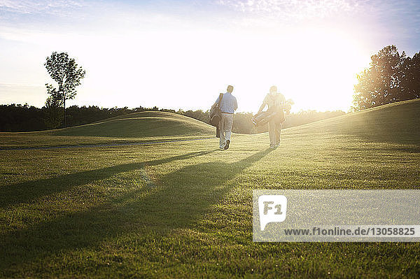 Rear view of golfers carrying bags while walking on field during sunset