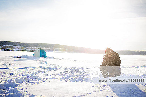 Mann sitzt bei Sonnenschein auf gefrorenem See gegen den Himmel