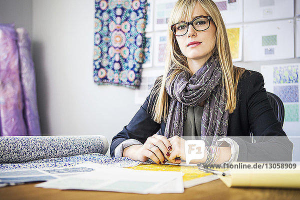 Portrait of confident female fashion designer sitting by desk at workshop