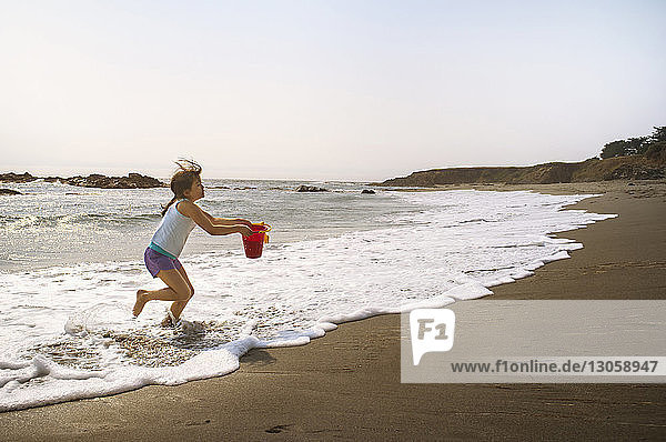 Side view of girl carrying bucket while running on shore at beach