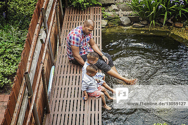 Hochwinkelaufnahme eines Vaters mit Söhnen  die auf einem Fußweg über einem Teich im Hinterhof sitzen