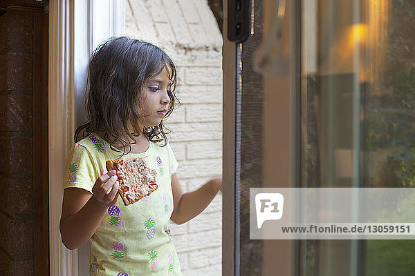 Nachdenkliches Mädchen hält Pizzastück in der Hand  während es zu Hause aus dem Fenster schaut