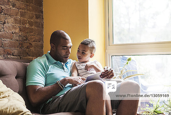 Mann benutzt Tablet-Computer  während er mit seinem Sohn zu Hause auf dem Sofa sitzt