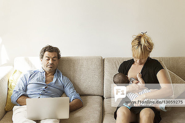 Mutter stillt ein Mädchen  während der Vater mit einem Laptop auf dem Sofa neben ihr sitzt