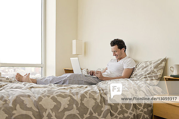 Side view of happy man using laptop on bed at home