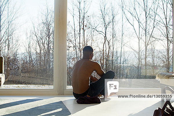 Rear view of man sitting on floor by window at home