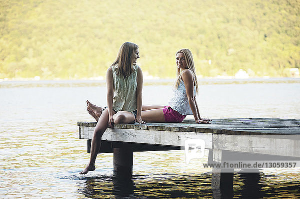 Lächelnde Freunde unterhalten sich  während sie auf dem Steg über dem See sitzen