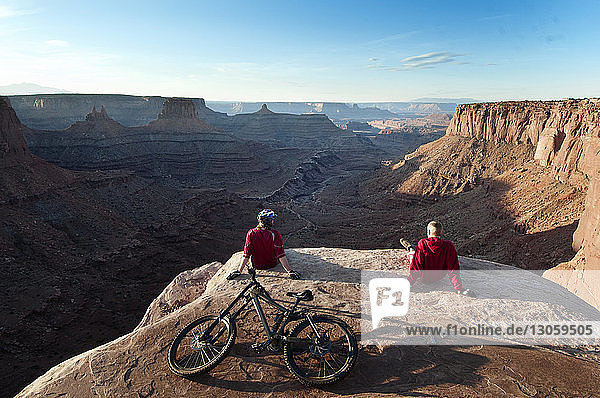 Rear view of mountain bikers relaxing on cliff against sky