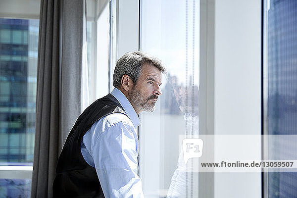 Nachdenklicher Geschäftsmann schaut durch Fenster  während er im Hotelzimmer steht