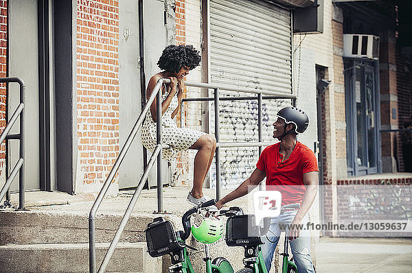 Mann mit Fahrrad schaut Frau an  die auf einem Geländer sitzt