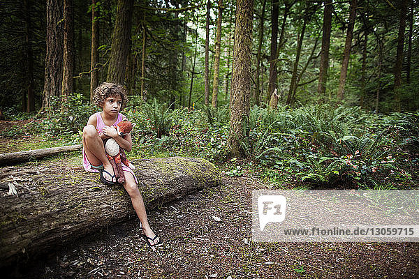 Trauriges Mädchen hält ausgestopftes Spielzeug in der Hand  während sie auf einem umgefallenen Baum im Wald sitzt