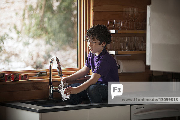 Junge füllt Wasser in Glas ein  während er an der Küchentheke sitzt