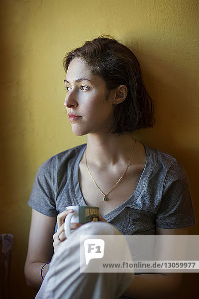 Close-up of thoughtful woman holding coffee cup and leaning on wall at home