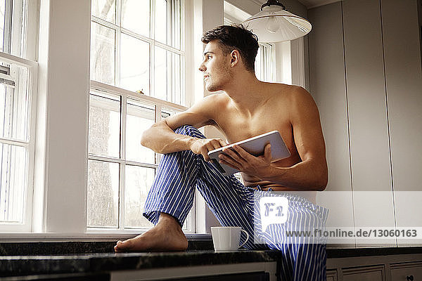 Mann mit Tablet-Computer schaut weg  während er auf der Küchentheke sitzt