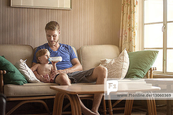 Lächelnder Vater schaut Baby Junge an  der zu Hause auf dem Sofa Milch trinkt