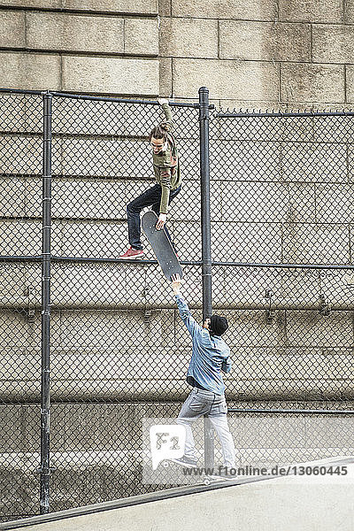 Man giving skateboard to friend hanging on chainlink fence against wall