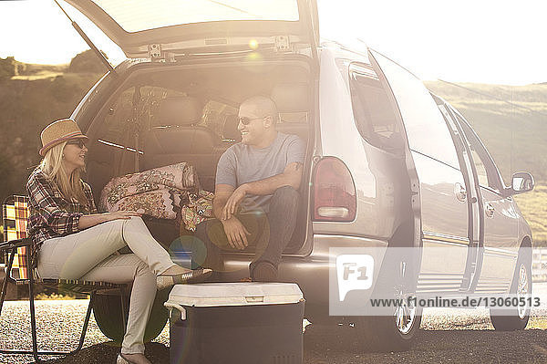 Man sitting in car trunk talking to woman on sunny day