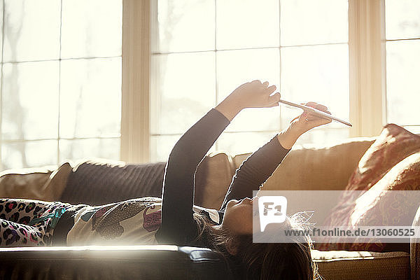 Side view of girl holding tablet computer while lying on sofa at home