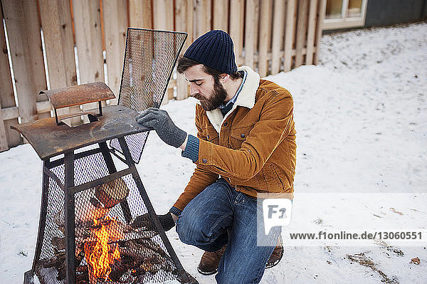 Schrägaufnahme eines Mannes,  der in einem schneebedeckten Hinterhof einen Baumstamm ins Lagerfeuer legt