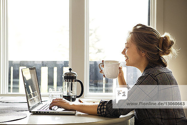 Glückliche Frau benutzt Laptop-Computer  während sie Kaffee am Tisch genießt